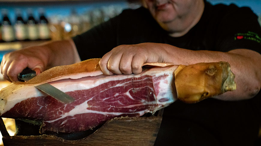 A chef carefully carves a thin slice from a large leg of cured prosciutto with a long, sharp knife.