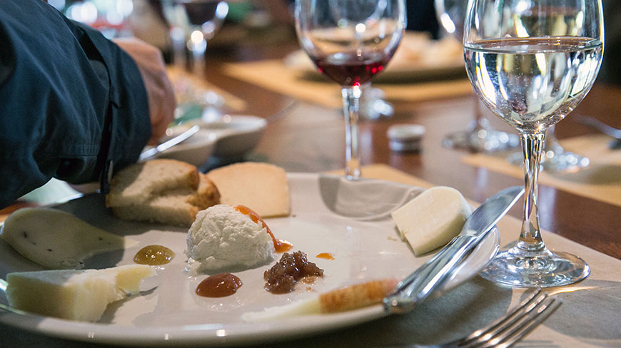 A close-up of a white plate with an assortment of cheeses and jams, next to glasses of wine and water on a table.