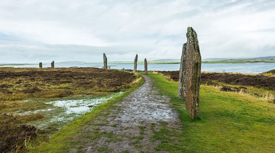 A dirt path curves alongside tall, weathered standing stones in a grassy field next to a large body of water.