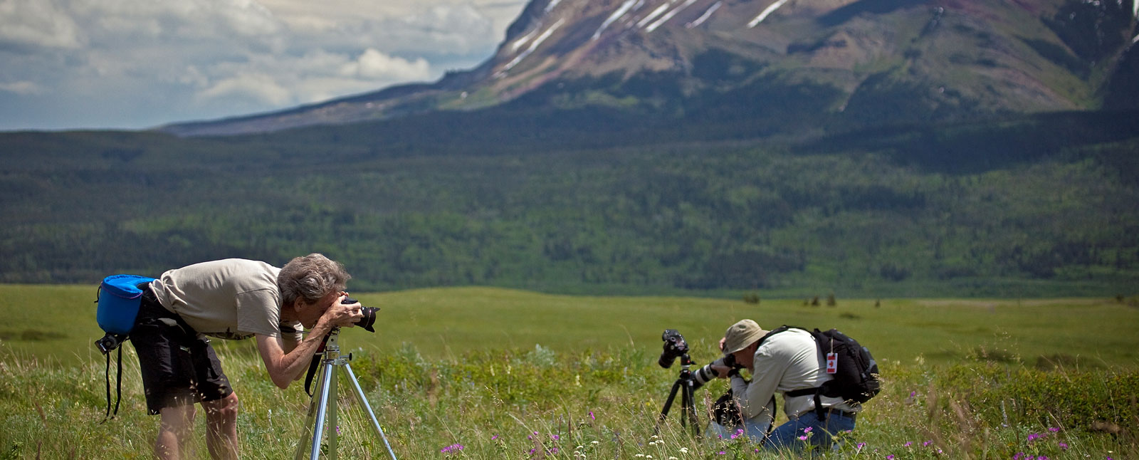 Two photographers with cameras on tripods take pictures in a vast green field at the base of a large mountain.