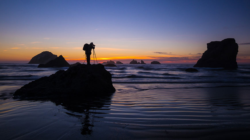A silhouetted photographer with a camera on a tripod captures a colorful sunset over a rocky coastline and the ocean.