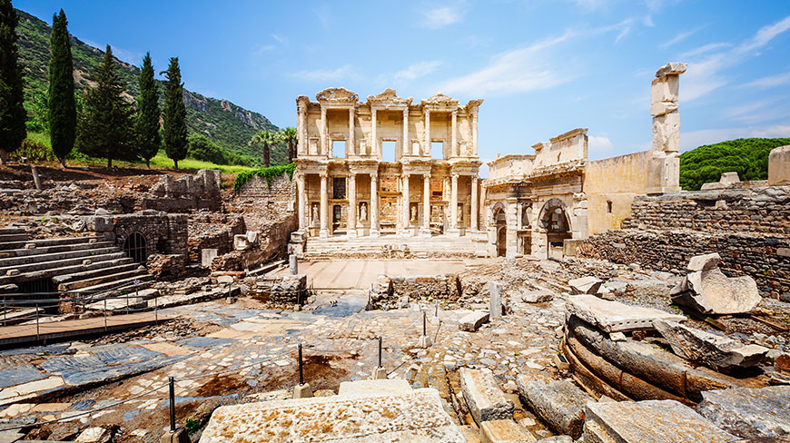 The ancient stone ruins of the Library of Celsus in Ephesus, Turkey, stand under a clear blue sky with hills in the background.