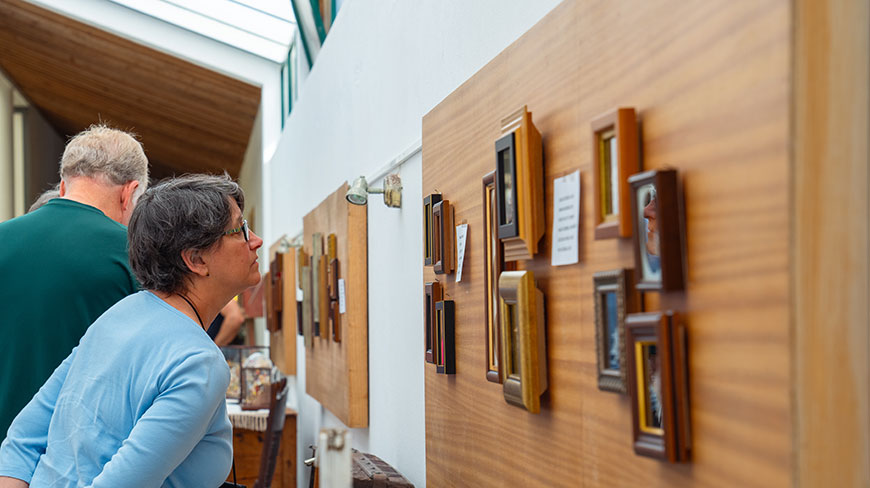 A woman in a blue shirt and glasses examines a wall of small, framed pictures in an art gallery or museum.