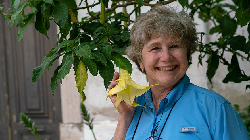 A smiling woman with gray hair in a blue shirt holds a large yellow trumpet-shaped flower next to a leafy plant.