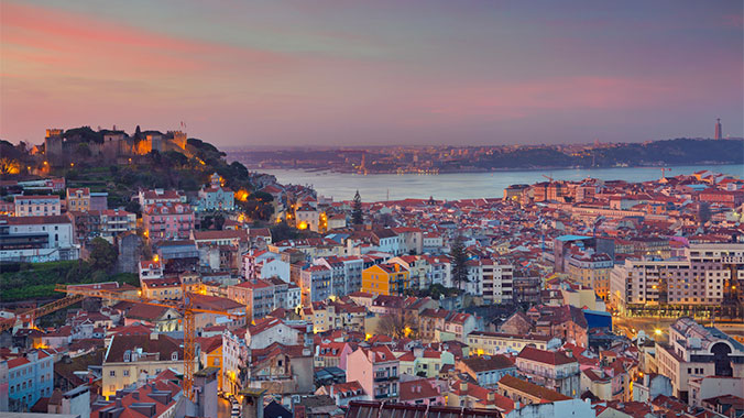 A sprawling cityscape of Lisbon, Portugal at sunset, with São Jorge Castle on a hill and the Tagus River in the background.