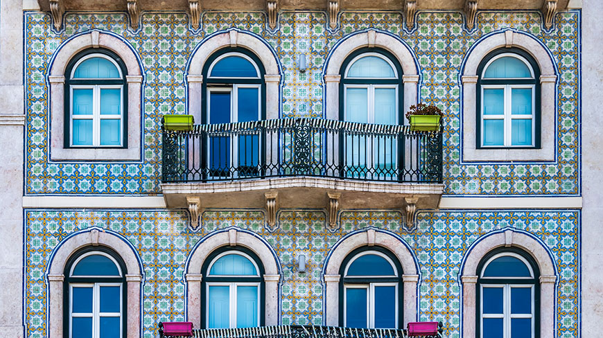 A building facade decorated with colorful patterned tiles, featuring arched windows and a small wrought-iron balcony.