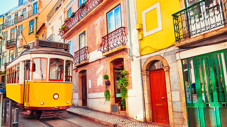 A classic yellow tram navigates a narrow cobblestone street lined with colorful, tiled buildings in Lisbon, Portugal.