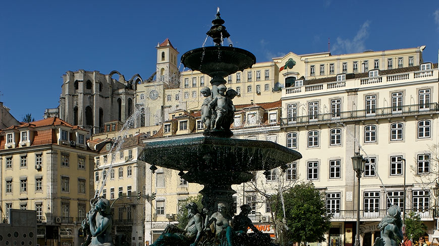 An ornate fountain in Rossio Square in Lisbon, with historic buildings and the Carmo Convent ruins in the background under a clear blue sky.