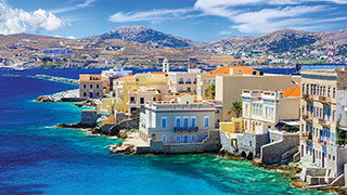 Colorful buildings on the Greek island of Syros line the rocky coast of the turquoise Aegean Sea under a partly cloudy sky.