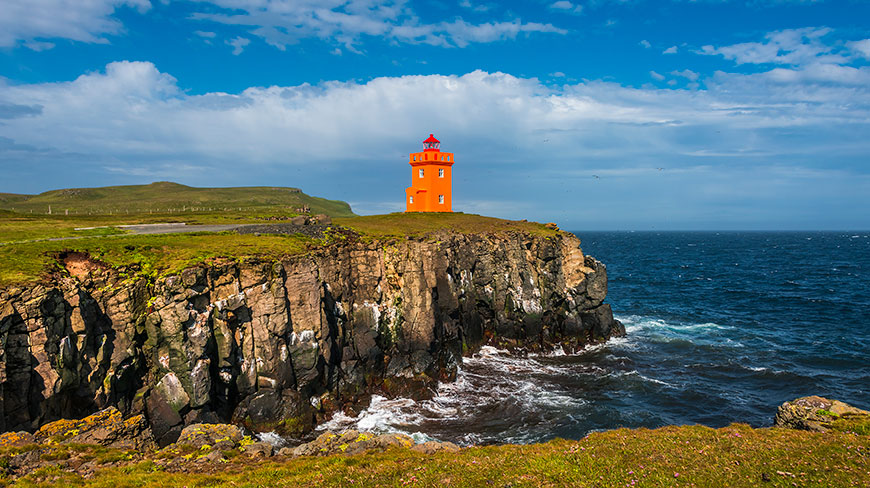 A bright orange lighthouse sits on a rugged cliff overlooking the ocean on Grímsey Island, Iceland, under a blue sky with fluffy white clouds.