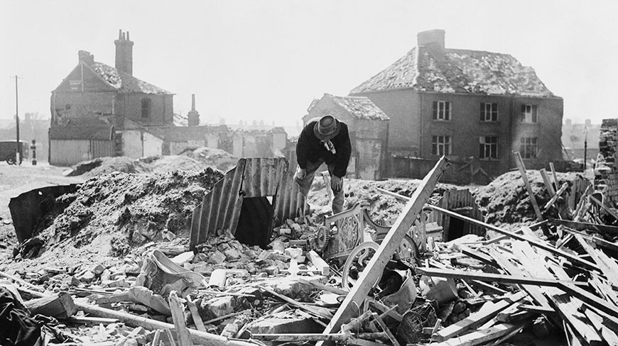 A man in a hat looks down at the rubble of bombed buildings in the UK during the Blitz of World War II.