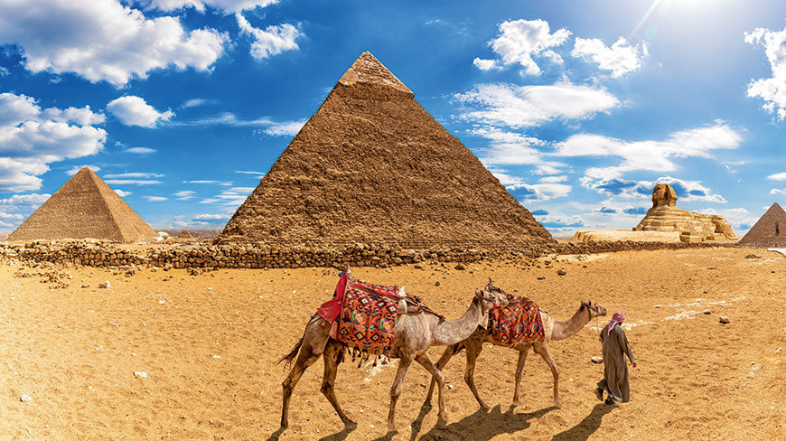 A person leads two camels through the desert in front of the Great Pyramids and Sphinx in Giza, Egypt.