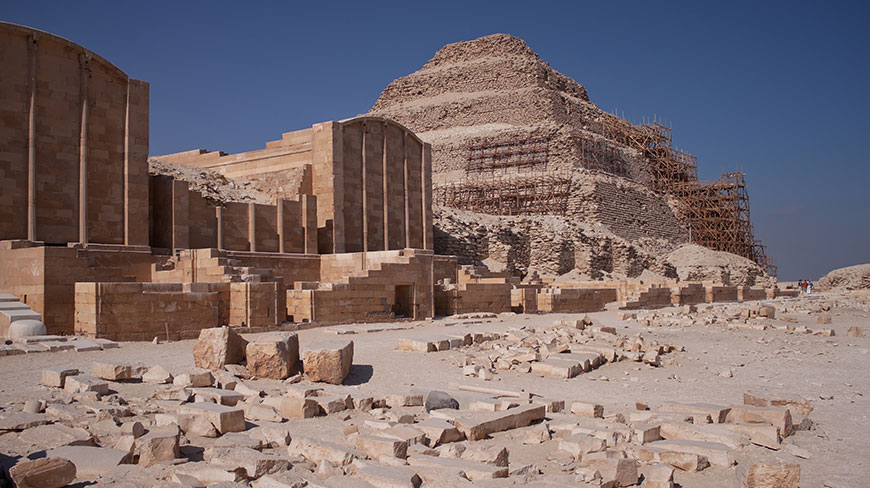 The Step Pyramid of Djoser at Saqqara, with stone ruins in the foreground and scaffolding on the pyramid for restoration.