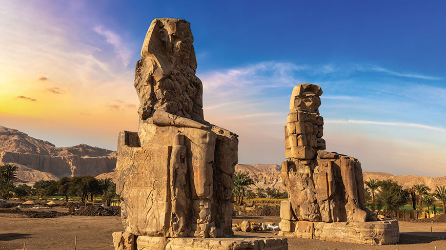 Two massive stone statues, the Colossi of Memnon, sit in a desert field in Luxor, Egypt against a colorful sunset sky and distant hills.