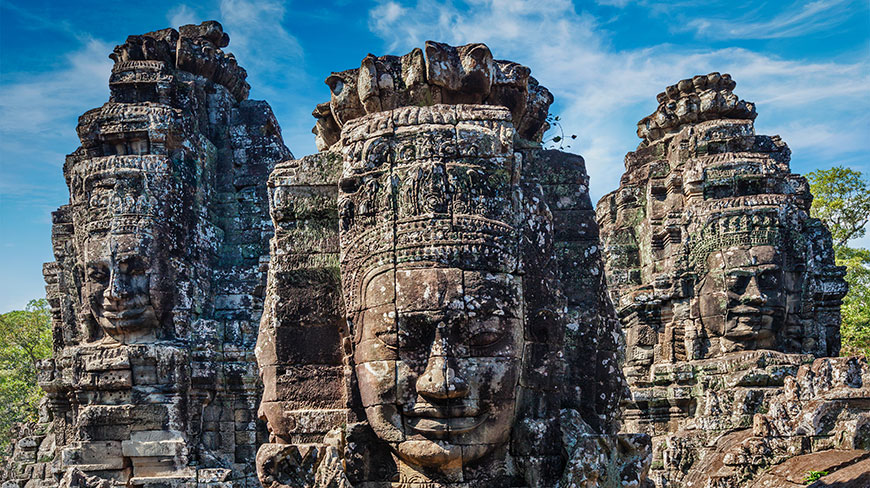 Three ancient, weathered stone towers at Angkor Wat in Cambodia feature large, serene faces carved into the rock under a blue sky.