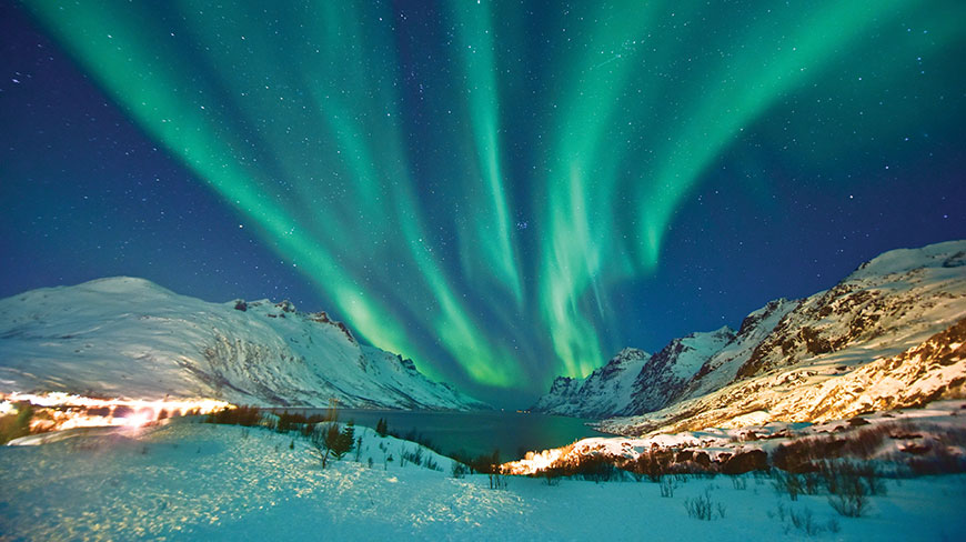The vibrant green aurora borealis glows in the starry night sky above snow-covered mountains and a fjord in Norway.