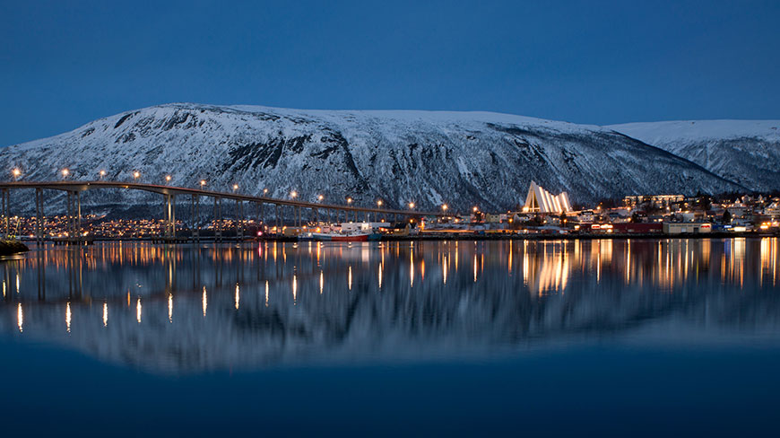 The city of Tromsø, Norway, is lit up at dusk with a bridge and the Arctic Cathedral reflected in the water before a snowy mountain.