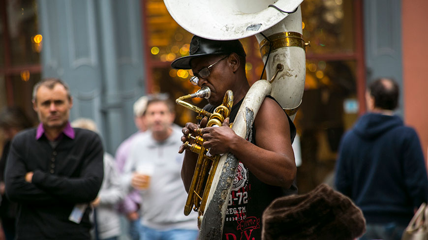 A musician plays a large brass sousaphone on a street in New Orleans, Louisiana, as people watch in the background.