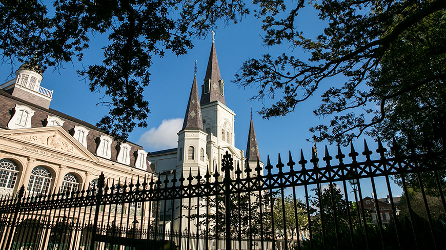 The Louisiana State Museum and St. Louis Cathedral in New Orleans are seen through the trees and behind an iron fence on a clear day.
