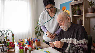 A female therapist assists an elderly man with a cognitive exercise at a table with art supplies.