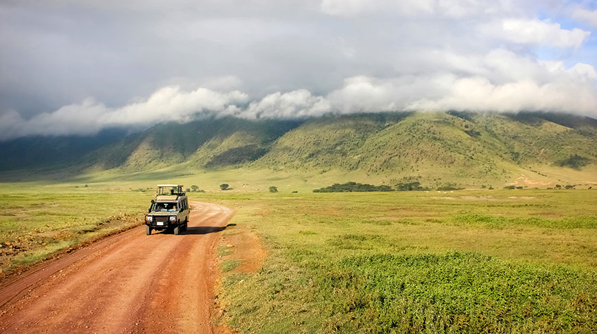 A safari vehicle drives on a dirt road across a grassy plain in Tanzania with the cloud-covered Ngorongoro Crater in the background.