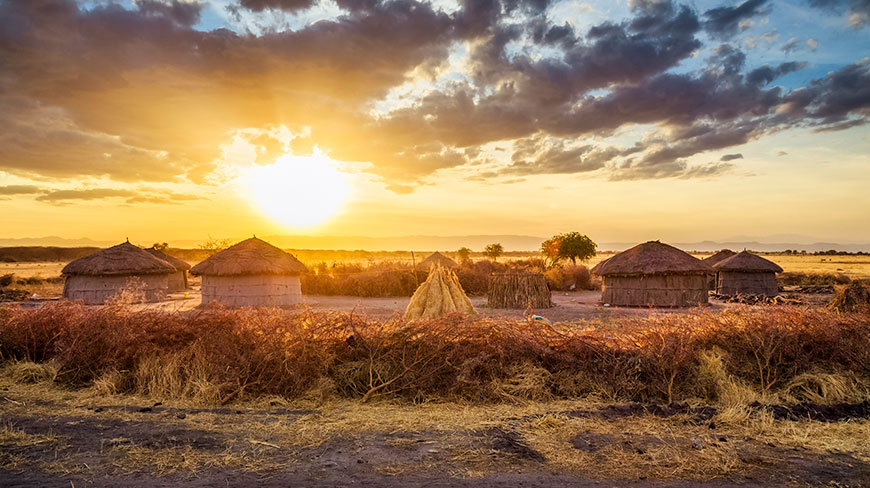 The sun sets over a Maasai village with traditional thatched-roof huts in Tanzania.