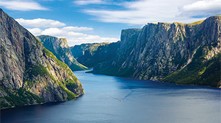 A boat sails through a fjord with steep, rocky cliffs under a blue sky in Gros Morne National Park, Newfoundland and Labrador.