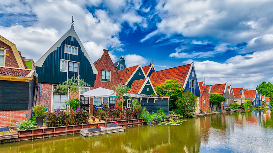 Colorful traditional Dutch houses with red-tiled roofs line a canal in De Rijp, Netherlands, under a blue sky with white clouds.