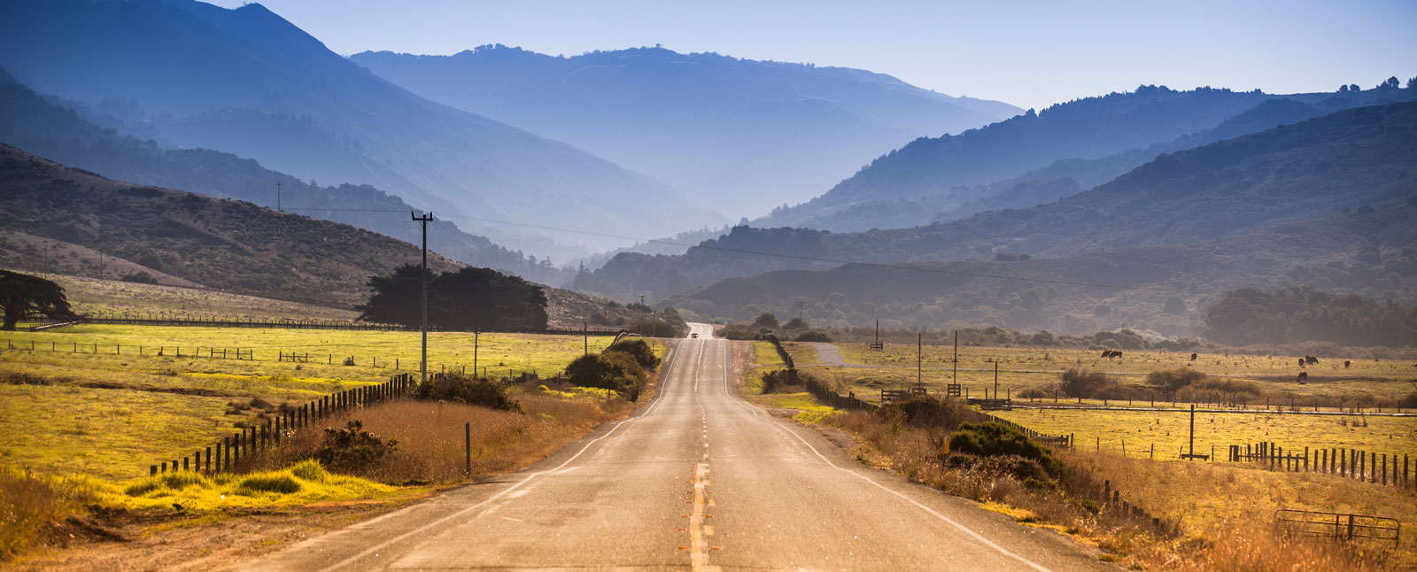 A scenic country road disappears into a hazy valley surrounded by green fields and layered blue mountains under a clear sky.
