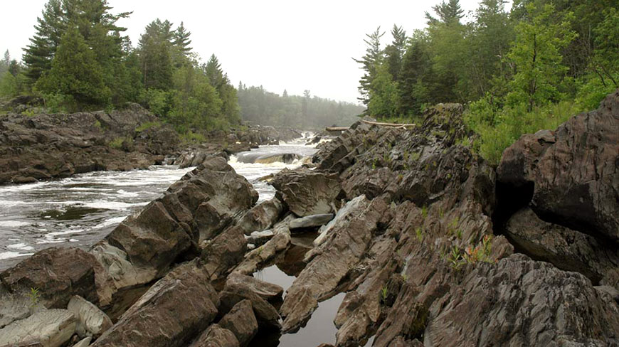 Tilted slabs of the Thomson Formation rock line the banks of a rushing river cutting through a forest in Wisconsin.