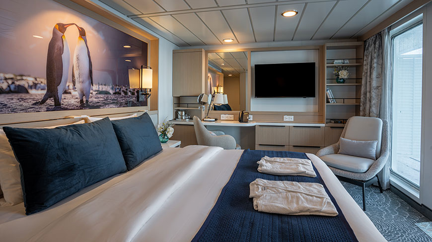 A well-lit stateroom on an Antarctic cruise ship, featuring a comfortable bed with blue accents and a large photograph of two penguins.