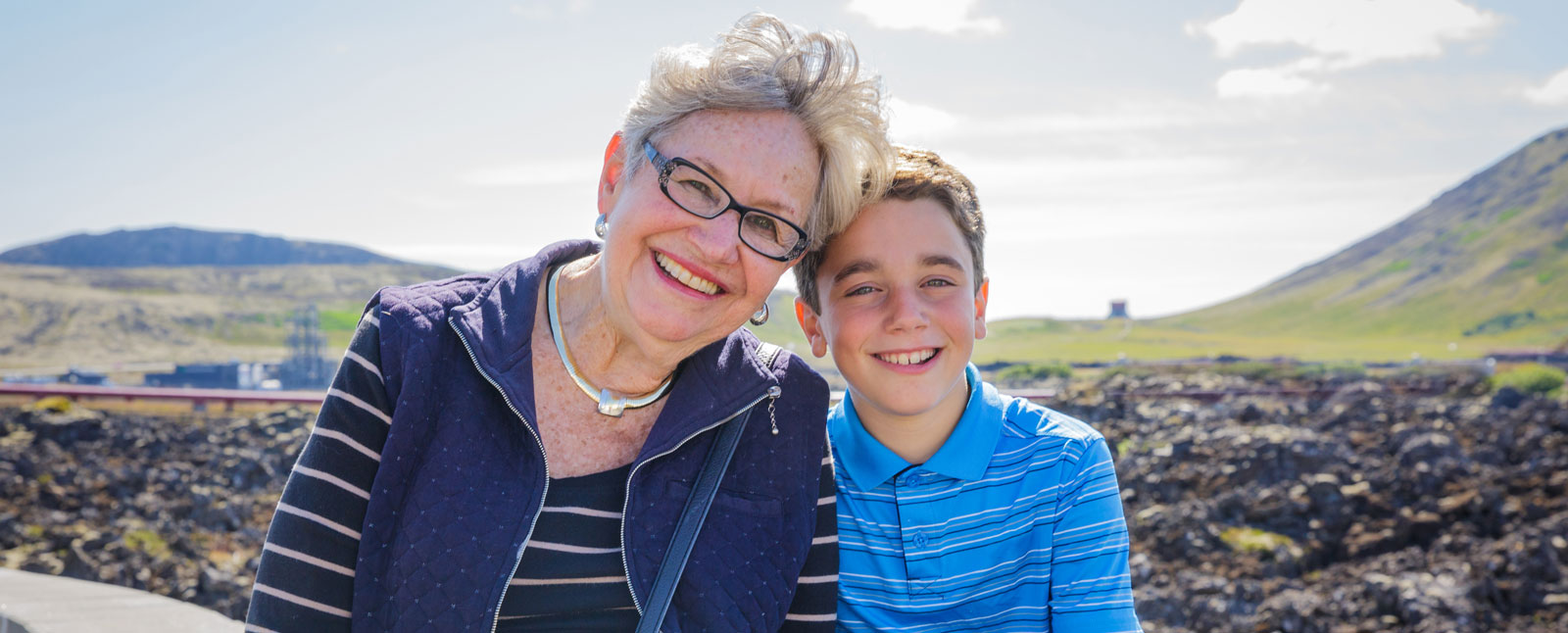 An older woman and a young boy smiling together outdoors in a rocky, volcanic landscape with mountains in the background.