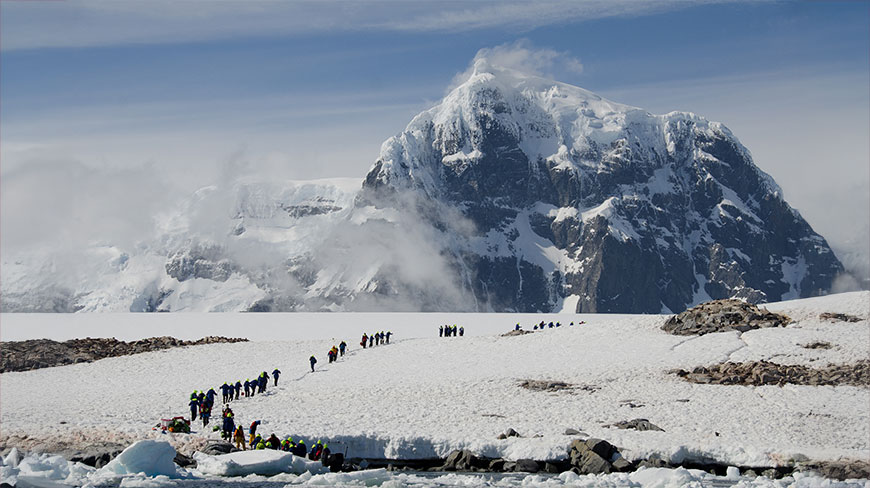 A group of explorers hike up a snowy landscape in Antarctica, with a large, cloud-wreathed mountain looming in the background.