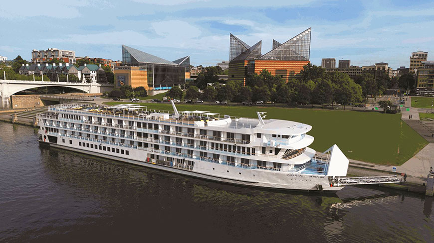 The American Serenade riverboat docked on the Mississippi River in front of a city with modern glass buildings under a blue sky.