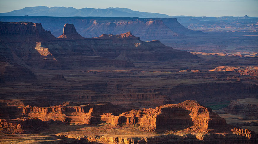 Golden sunlight illuminates the mesas and buttes of a vast, layered canyon landscape with distant blue mountains on the horizon.