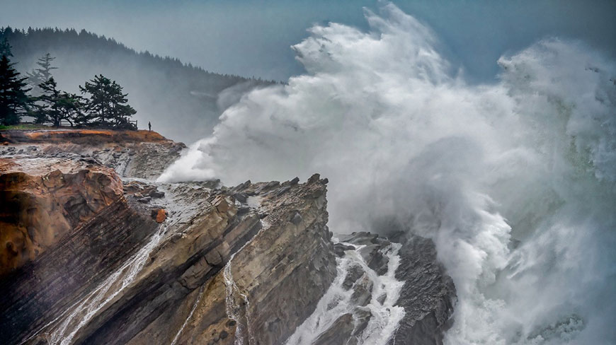 A person stands on a rocky cliff watching enormous white waves crash against the shore, with a forested hillside in the background.