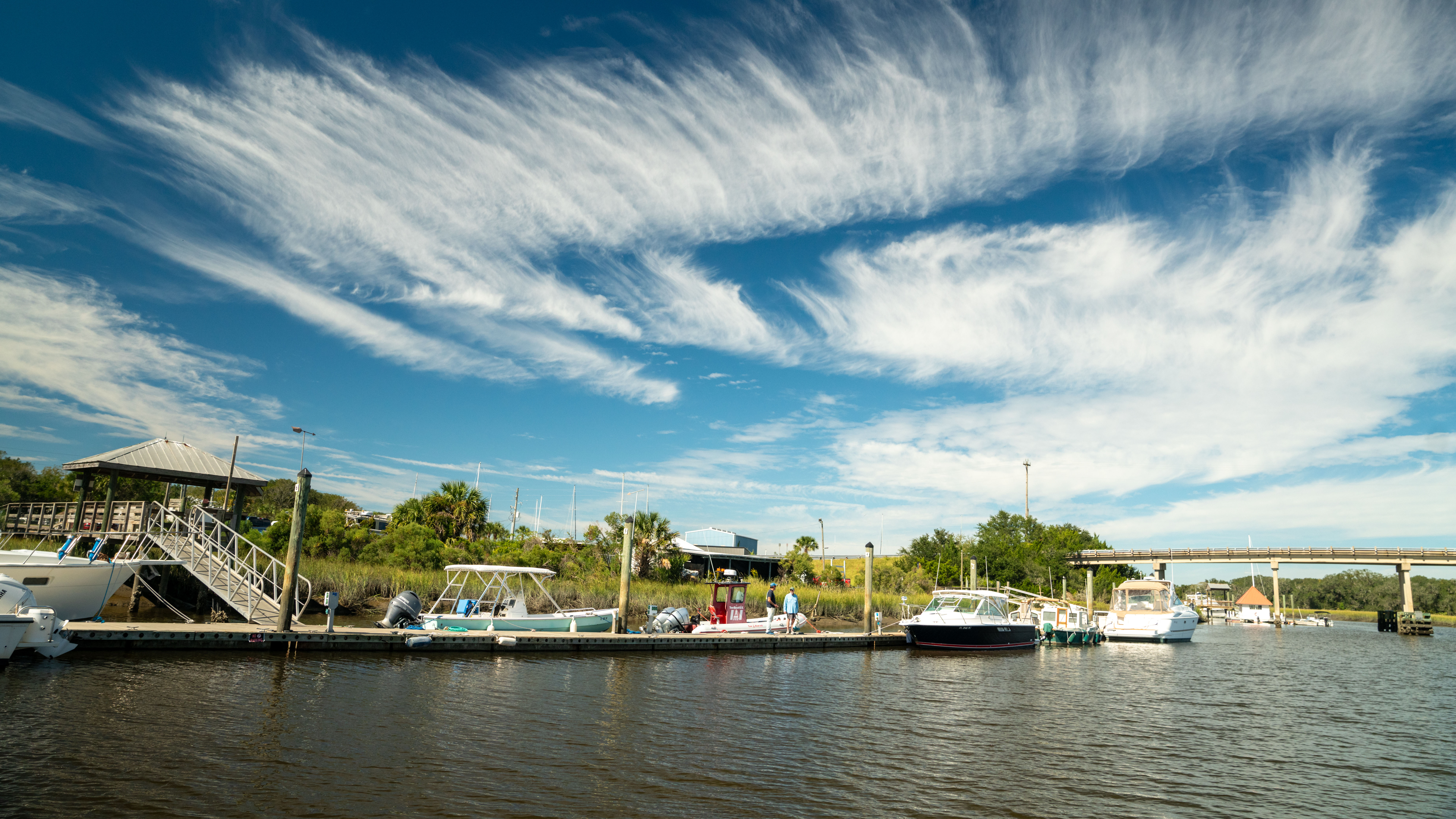 Boats are docked at a marina on a sunny day in Florida, under a blue sky with sweeping white clouds.