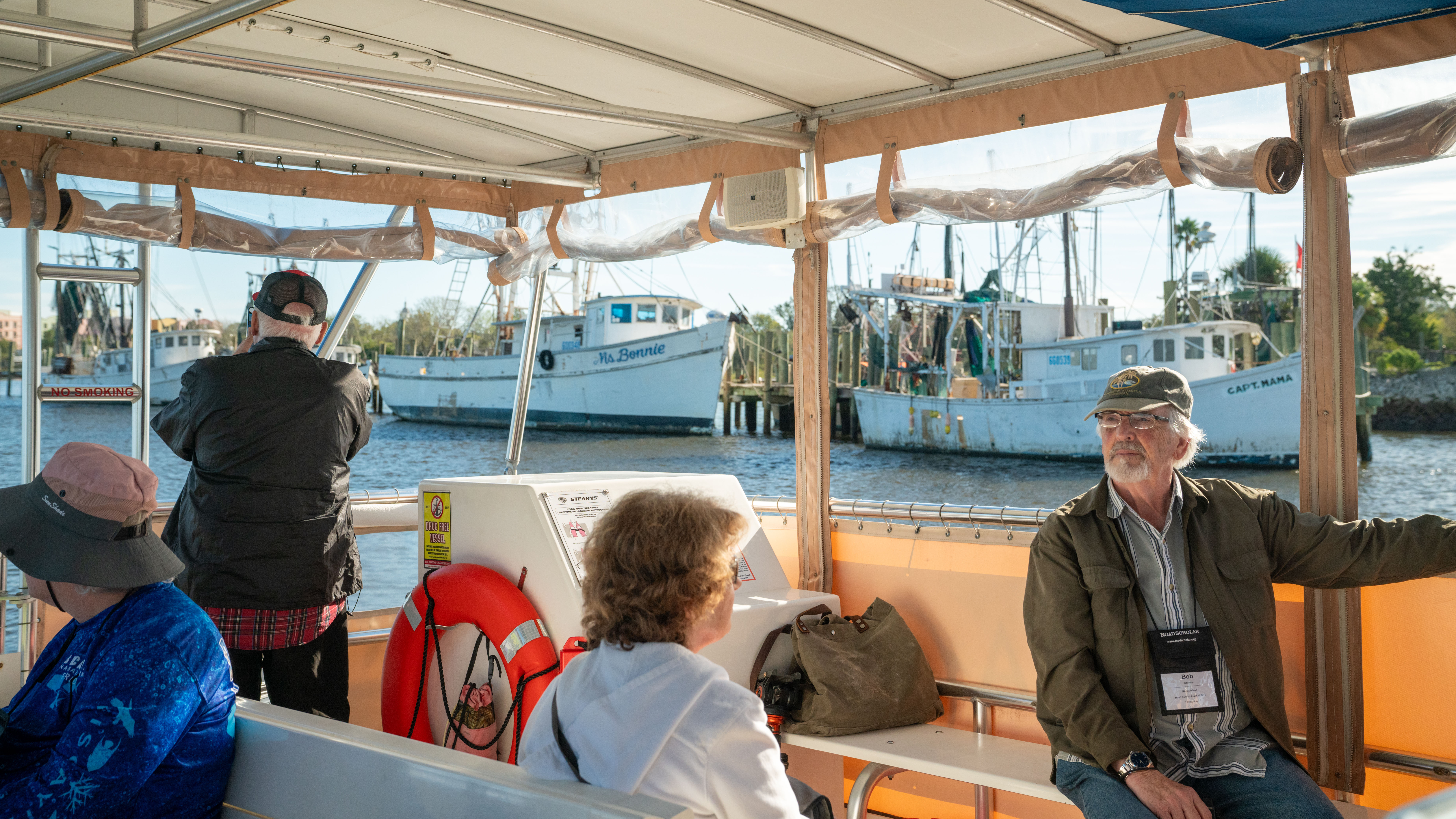 A group of passengers on a covered tour boat cruise along the water past docked fishing boats on a sunny day in Florida.