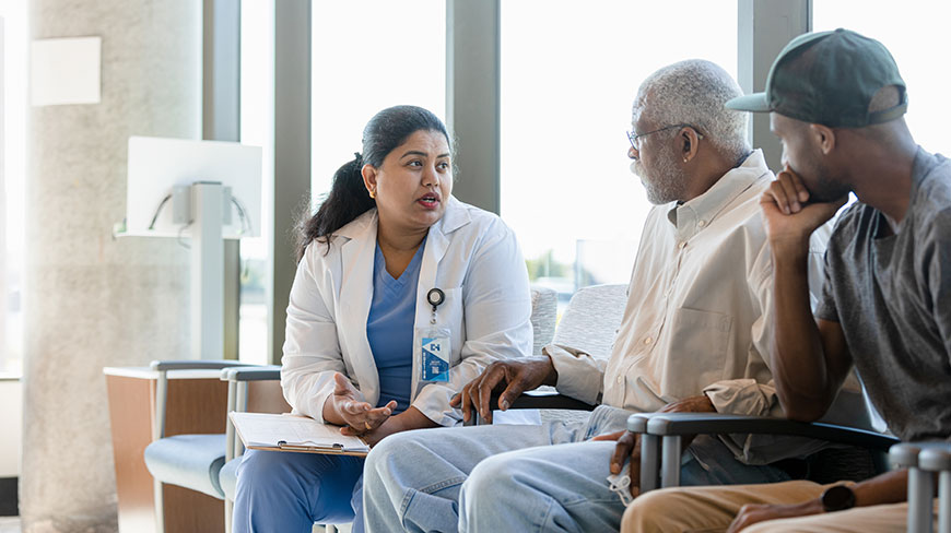 A female doctor sits and talks with an older male patient and a younger man in a clinic waiting room.