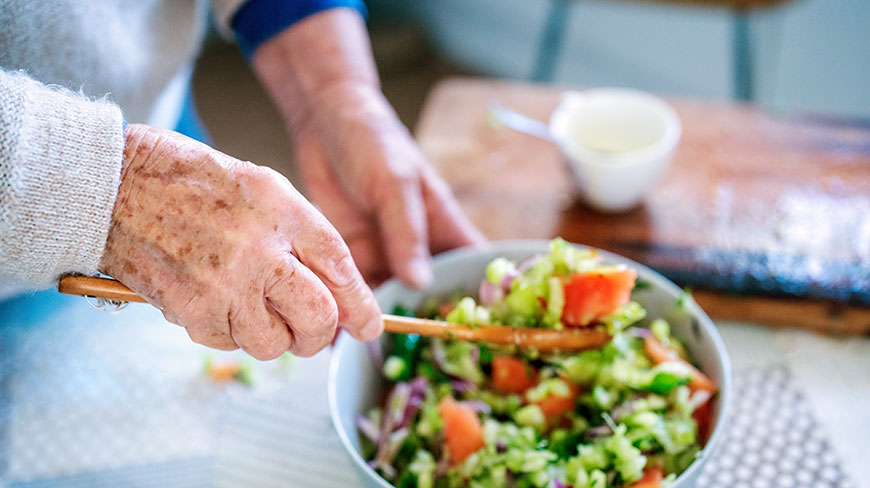 An older person's hands toss a fresh, colorful salad in a bowl with wooden spoons.