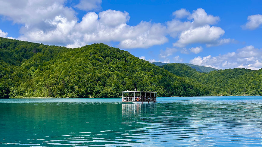 A tour boat carries passengers across a turquoise lake surrounded by lush green hills in Plitvice Lakes National Park, Croatia.