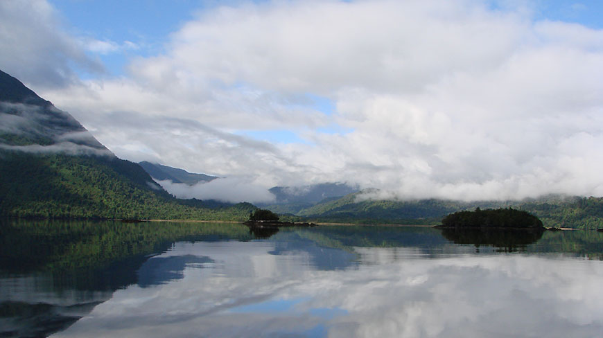 A calm fjord in Chile/Argentina with green mountains and clouds reflected in the still water.