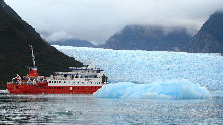The red and white Skorpios II cruise ship floats near the massive San Rafael Glacier and a large iceberg in the waters of Chile.