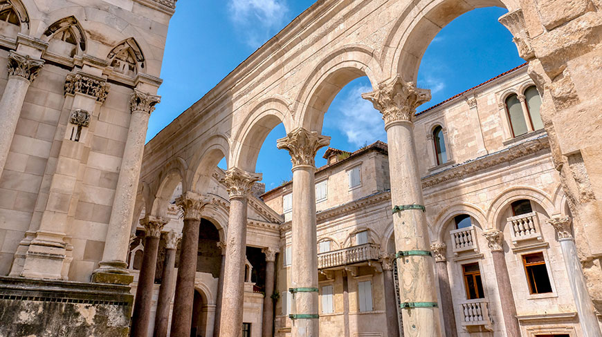 Sunlight illuminates the ancient stone columns and arches of the Peristyle in Croatia under a clear blue sky.