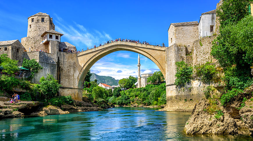 The historic Stari Most stone bridge arches over a turquoise river in a town surrounded by lush greenery under a bright blue sky.