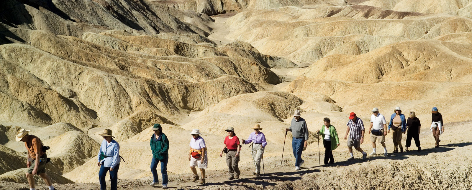 A small group of hikers follows a trail through a vast, arid landscape of golden, rolling hills under a clear blue sky.