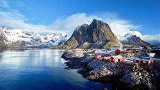 Red fishing cabins sit on the rocky coast of Reine in Norway's Lofoten Islands, with snow-covered mountains rising from the sea.