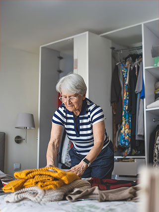 An older woman with white hair and glasses folds a pile of sweaters on a bed in front of an open closet.