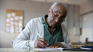 An older Black man with glasses wearing a grey cardigan over a green shirt, writing in a notebook at a desk.