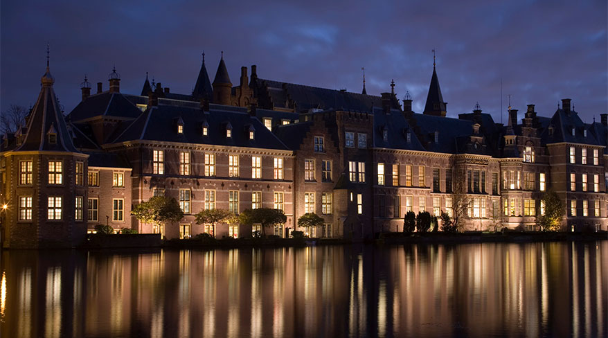 A historic building complex in Amsterdam, Netherlands, is illuminated at night, reflecting in the adjacent calm water.